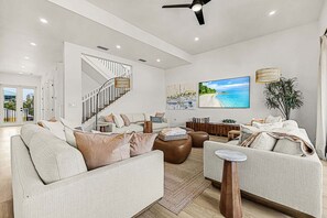 A modern living room with a staircase, comfortable seating, a flat-screen TV, and a large window. - 163 Tranquility Tides : 6br W/ Pool And Spa! 6 Bedroom Home (Fort Myers Beach)