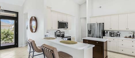 A modern kitchen with a central island, bar stools, a microwave, a stove, a refrigerator, and a coffee maker.
