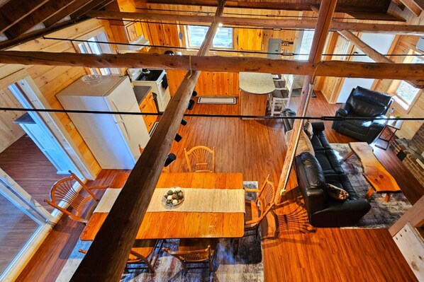 A spacious loft-style room with a high ceiling, wooden beams, a dining area with a table and chairs, a black leather sofa, and a view of the outdoors.