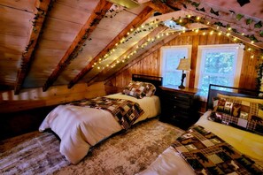 Une chambre mansardée chaleureuse avec un plafond en pente, des murs en bois et des guirlandes lumineuses. On y trouve deux lits avec des couvertures et une table de chevet avec une lampe.