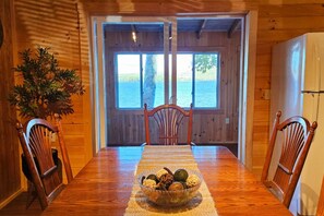 Une table à manger en bois, entourée de chaises, un saladier rempli de fruits et, par la porte, une vue sur l’eau.
