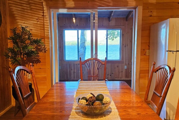 Una mesa de comedor de madera con sillas, un cuenco de fruta y vista al agua a través de una puerta.