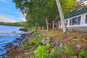 A lakeside cabin with a wooden deck, a stone wall, and a dock extending into the water. - The Water's Edge | Peaceful Lakeside Cabin (Hartland)