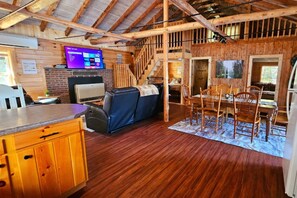 A rustic cabin interior with a wooden dining area, a sofa, and a TV mounted on the wall.