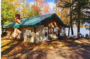 Une cabane rustique avec un toit végétalisé, des fondations en pierre et une petite véranda équipée d’une table et de chaises.