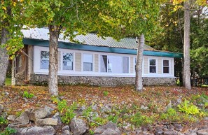 Eine rustikale Holzhütte mit Steinfundament, weißer Fassade und einem grünen Dach. Bäume und felsiger Boden mit etwas Grün umgeben.