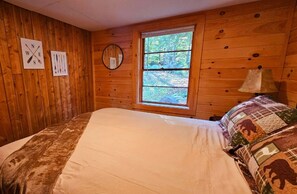 Ein Schlafzimmer einer Holzhütte mit einem großen Bett, einem Fenster mit Blick auf den Wald und Wanddekorationen, darunter ein Kompass und ein runder Spiegel.