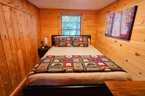 Un dormitorio acogedor de cabaña de madera con una cama, una mesita de noche, una ventana y un cuadro en la pared.