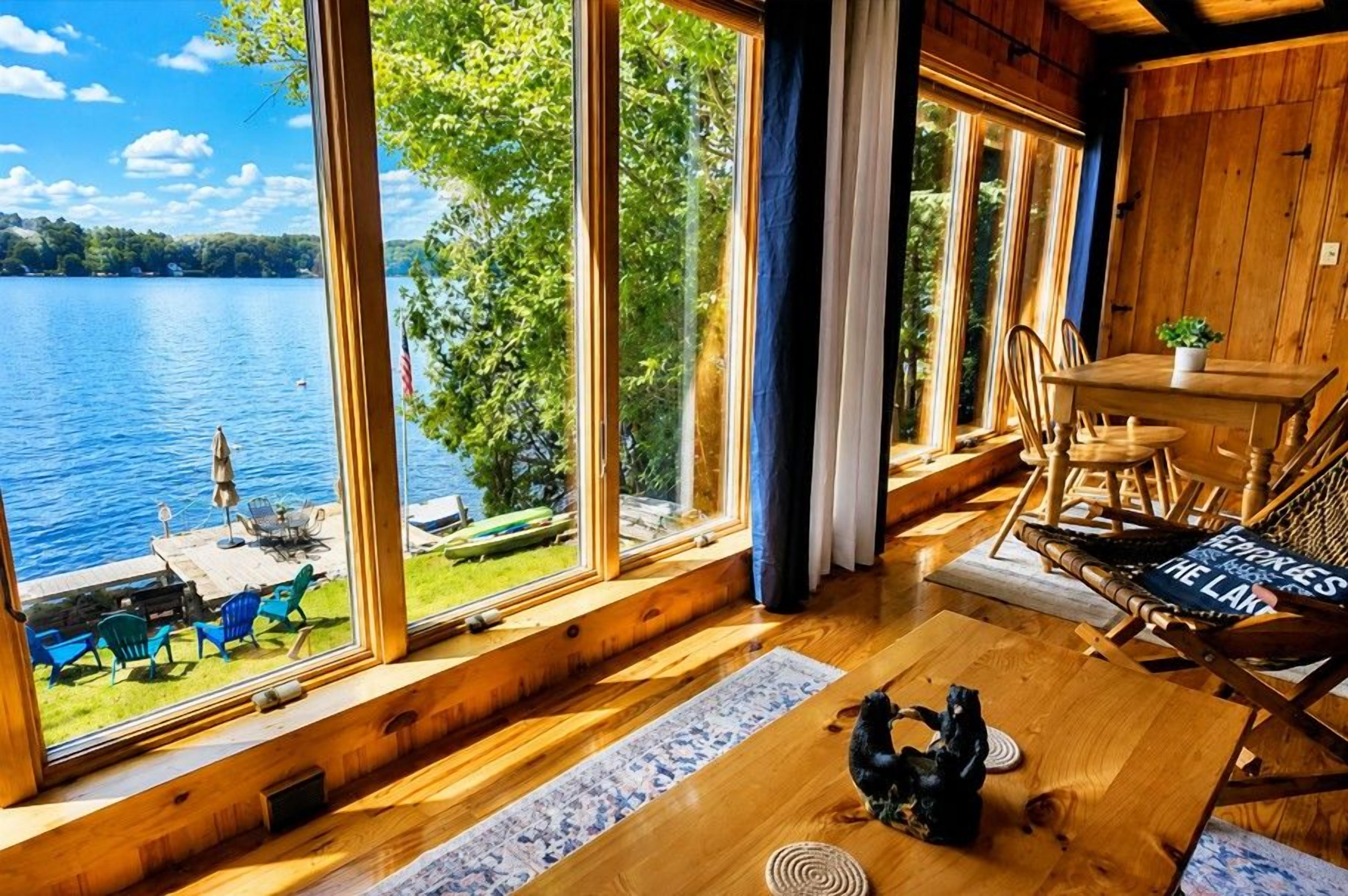 A cabin with large windows overlooking a lake, a dining area with a table and chairs, and a cozy seating area with a rug and a decorative item.