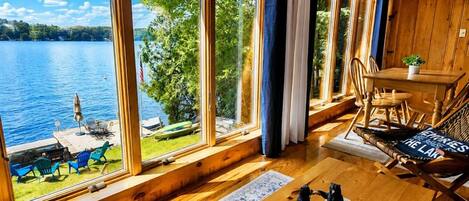 A cabin with large windows overlooking a lake, a dining area with a table and chairs, and a cozy seating area with a rug and a decorative item.