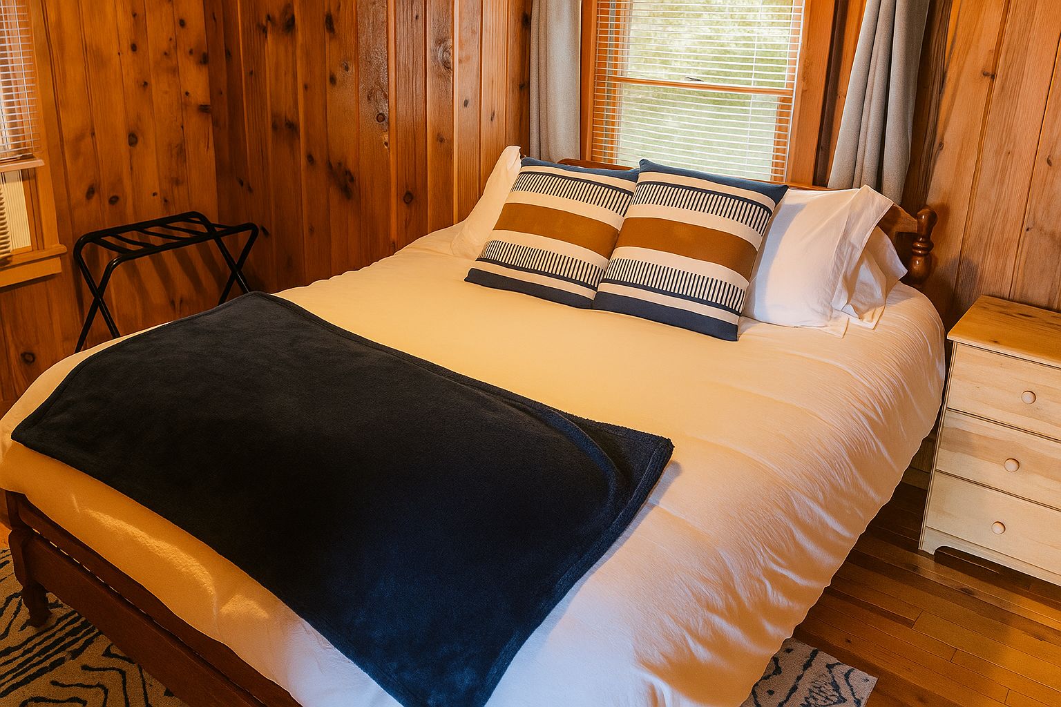Una cama bien hecha con un plaid oscuro, una mesita de noche de madera con cajones y una silla frente a una pared de madera con una ventana.