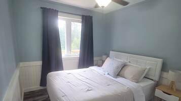 A bedroom with a bed, a ceiling fan, a window with curtains, and a nightstand with a lamp.