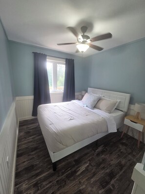 A bedroom with a bed, a ceiling fan, a window with curtains, and a nightstand with a lamp.