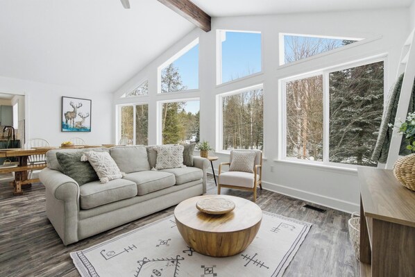 A spacious living room with a high ceiling, large windows, a grey sofa, a round wooden coffee table, and a dining area with a wooden table and chairs.