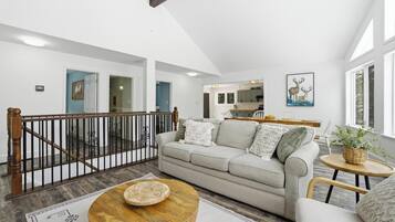A spacious living area with a high ceiling, a staircase, a kitchen in the background, and a large window.
