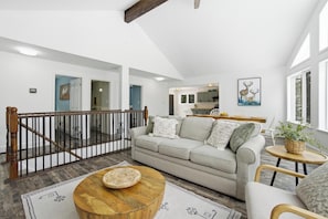 Un espacio habitable espacioso con techo alto, una escalera, una cocina al fondo y un ventanal grande.