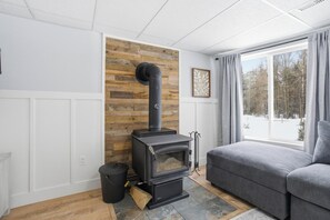 A modern living room with a wood-burning stove, a grey sofa, and a window with a view of a snowy landscape.
