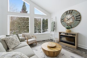 A cozy living room with a large window, a wooden coffee table, and a wall clock.