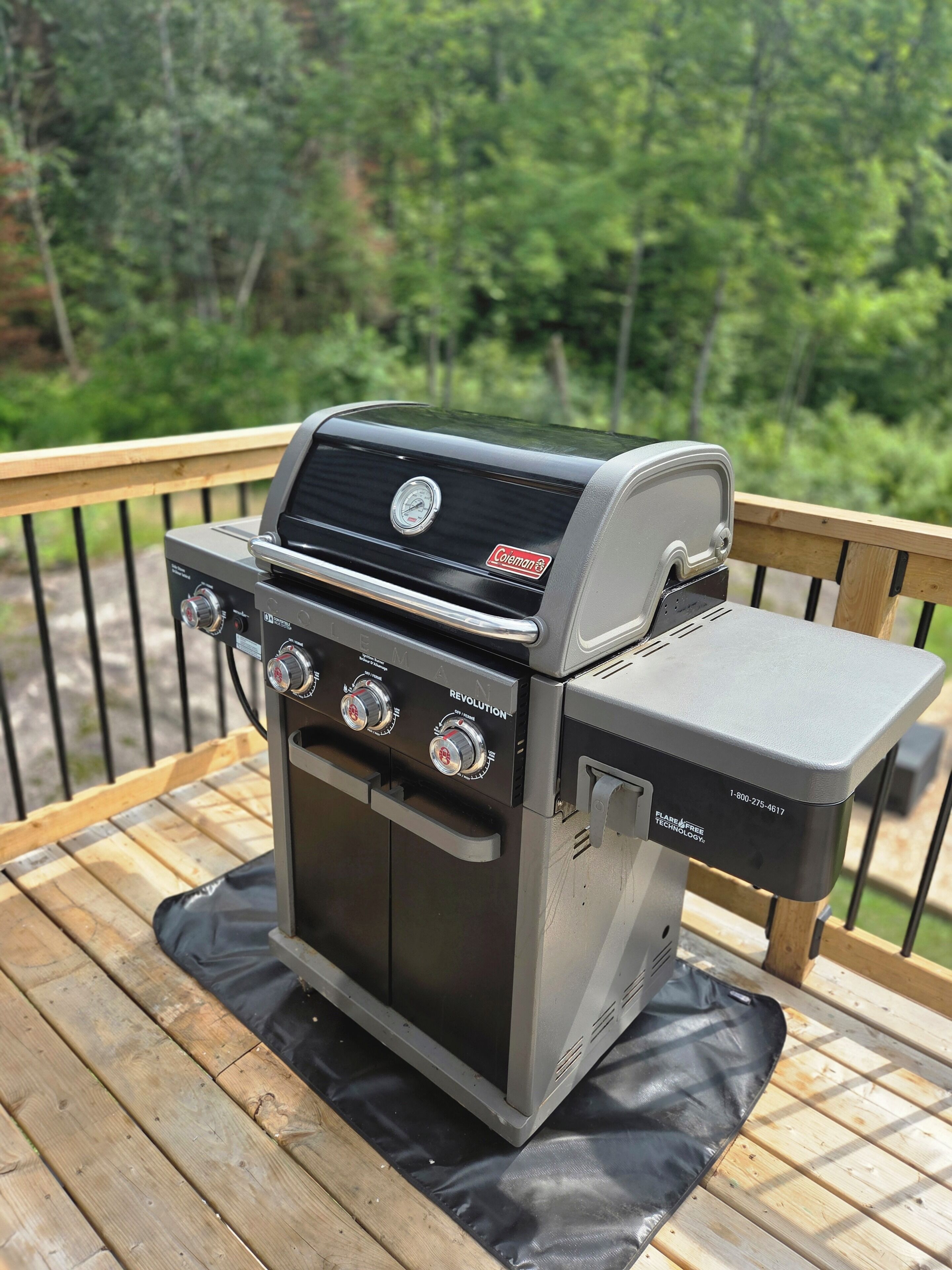 A Coleman gas grill with four burners, a digital display, and a side shelf, placed on a wooden deck with a metal railing.