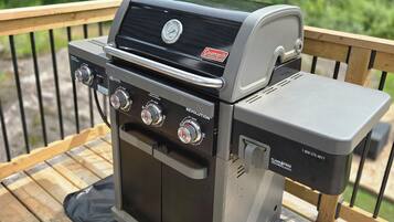 A Coleman gas grill with four burners, a digital display, and a side shelf, placed on a wooden deck with a metal railing.