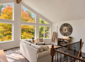 A spacious living room with a large window overlooking a forest, a cozy sofa, a wooden staircase, and a rustic wall clock.