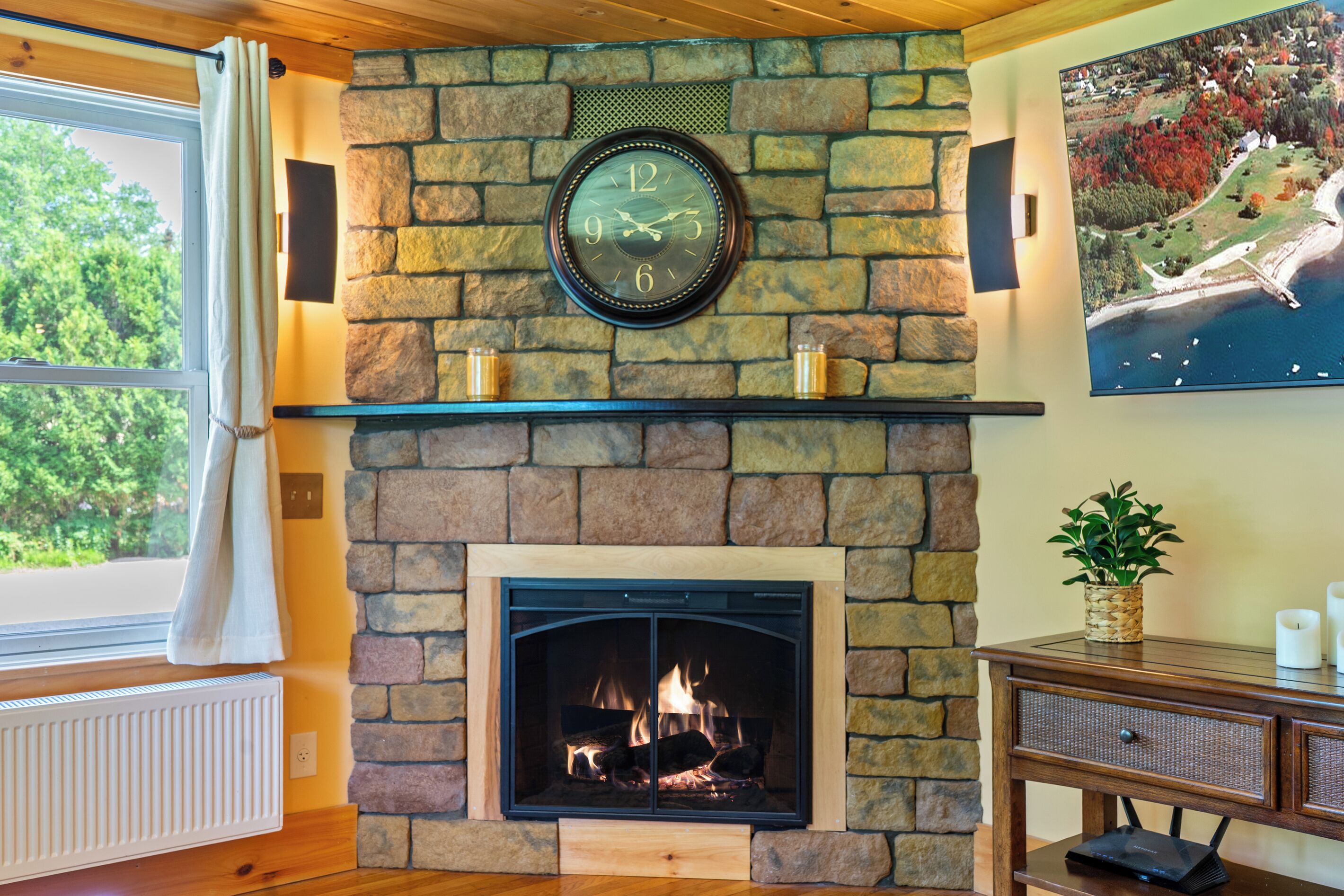 A stone fireplace with a clock above it, a window with curtains, and a framed picture on the wall.