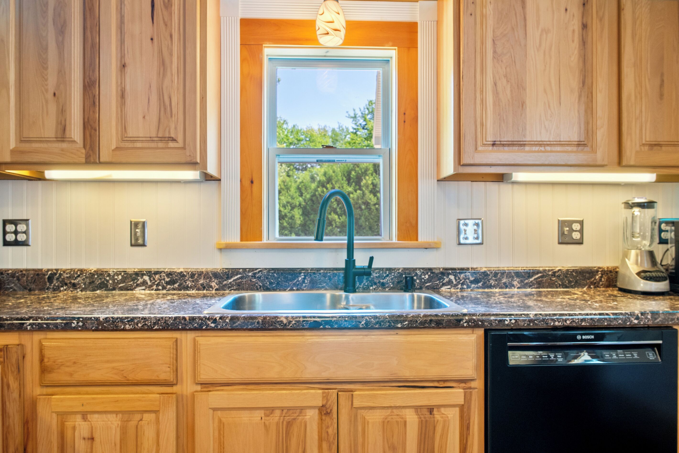 A kitchen with wooden cabinets, a stainless steel sink, a Bosch dishwasher, and a window with a view of greenery.