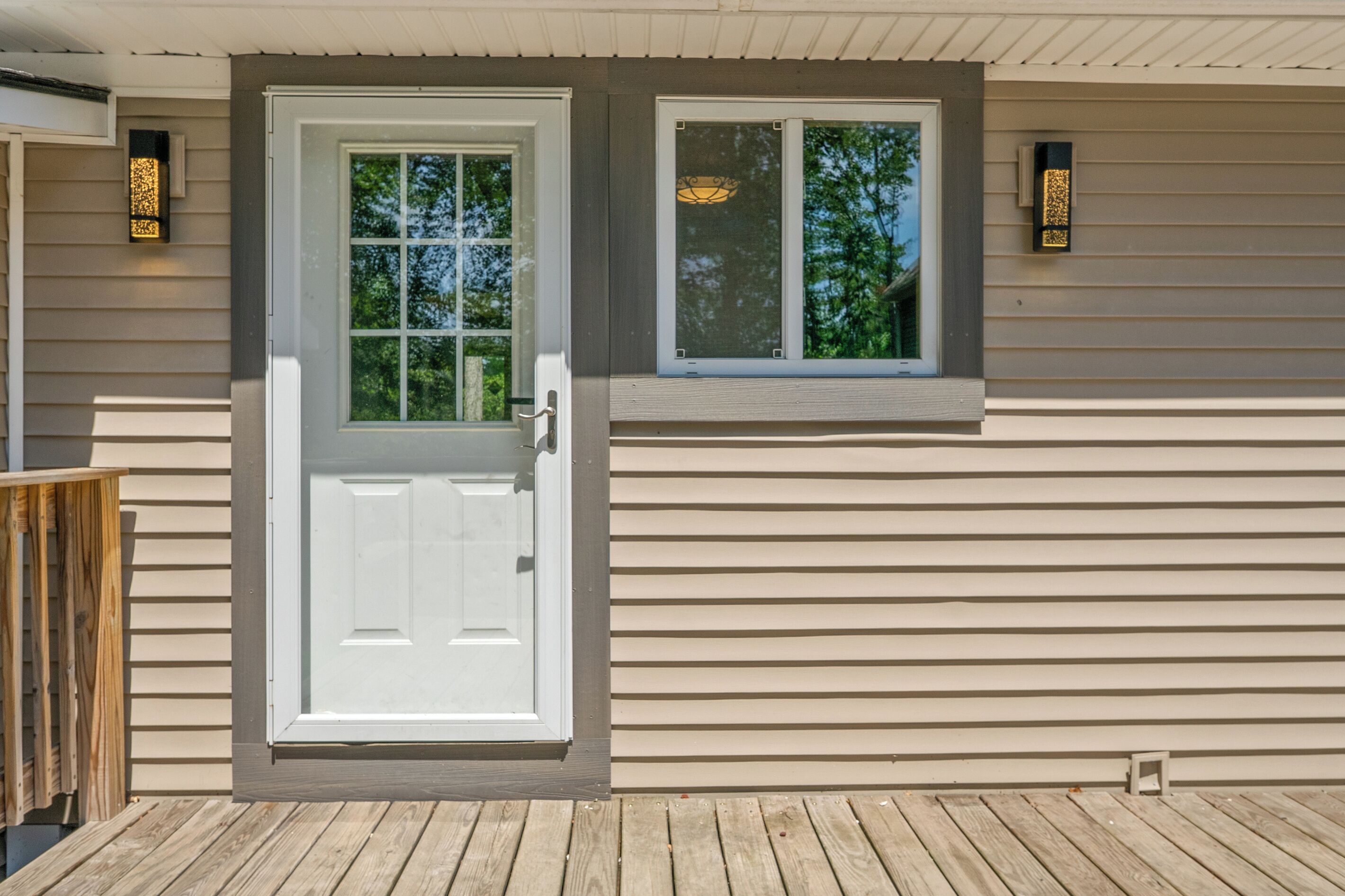 A house with a white front door, a small window with a view of trees, and two wall-mounted light fixtures.