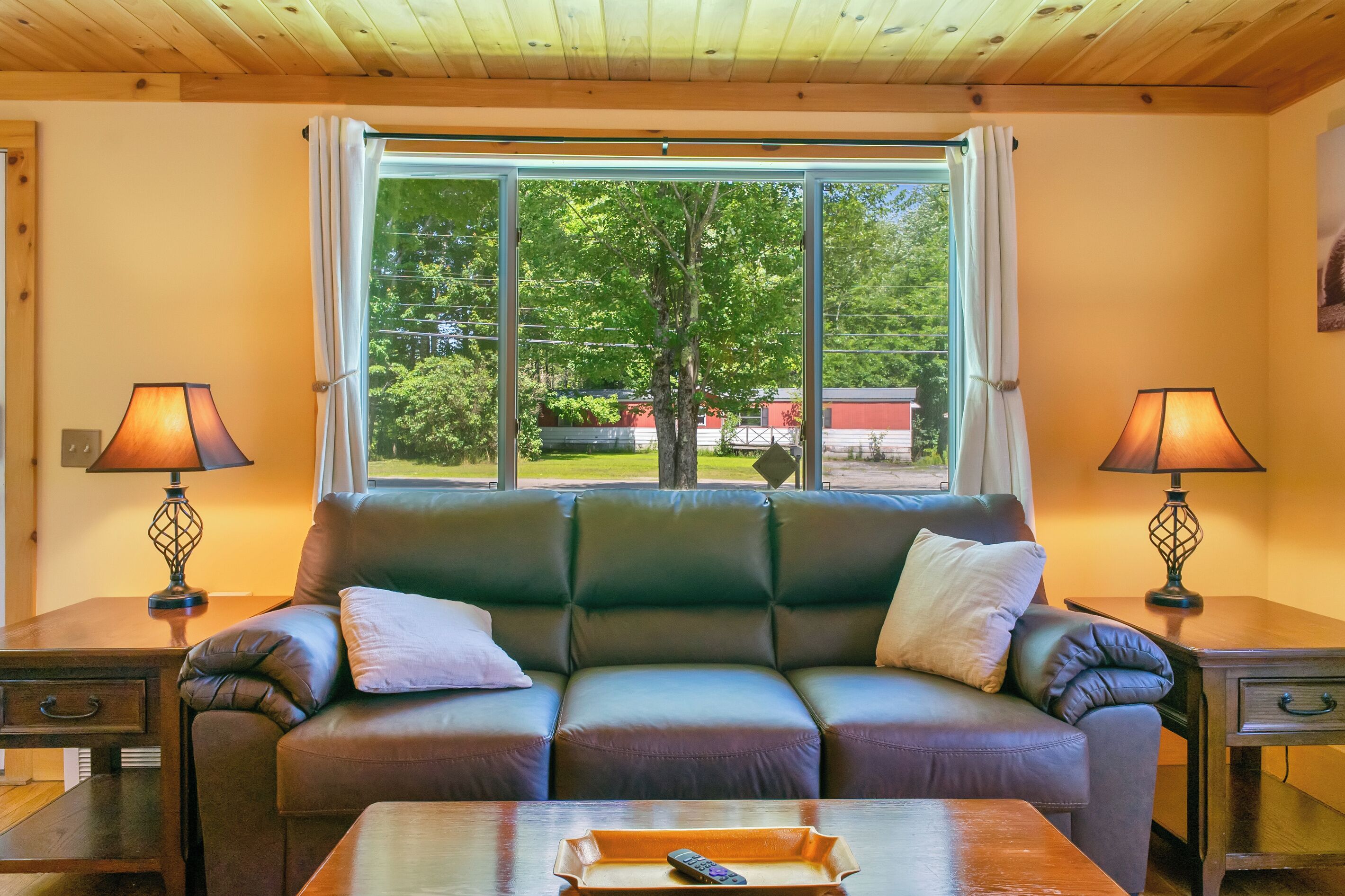 A cozy living room with a leather sofa, two lamps, a wooden coffee table, and a large window with a view of trees and buildings.