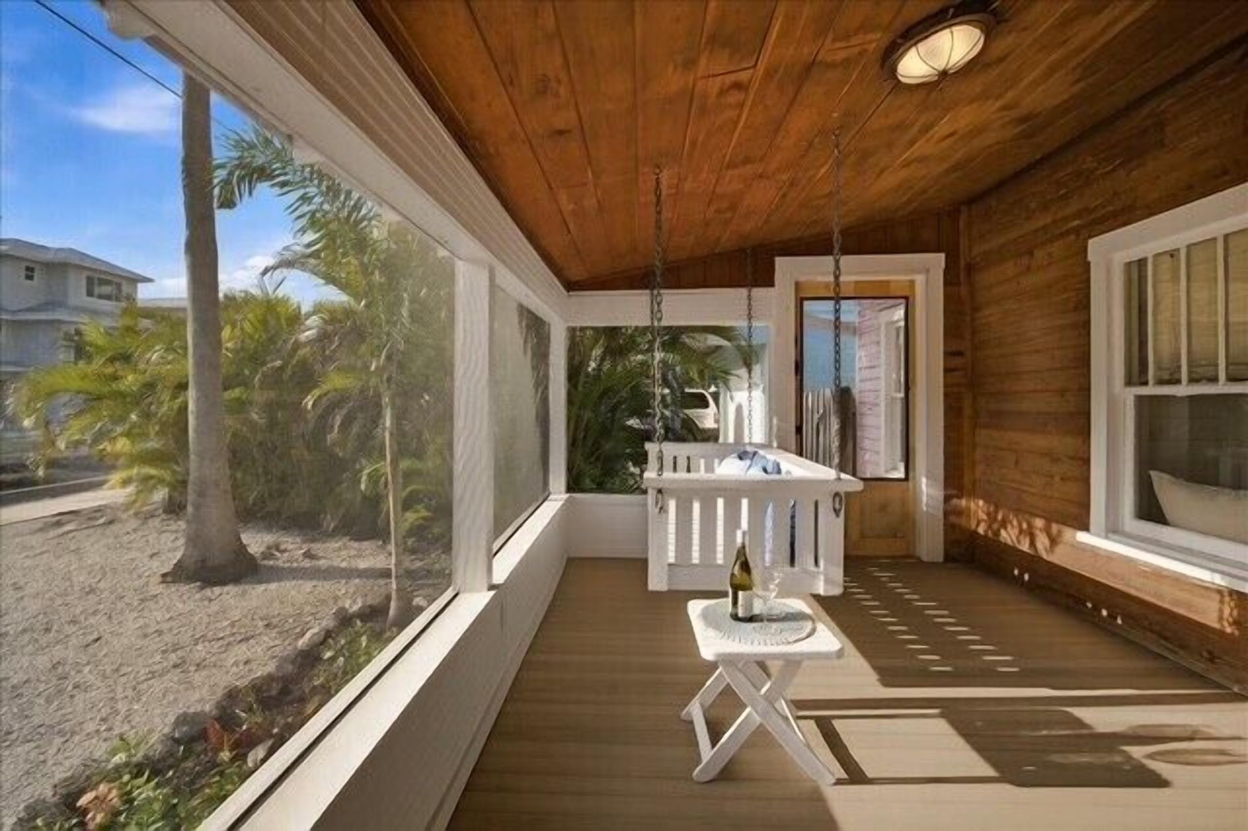 Une terrasse en bois avec une balustrade blanche, une petite table avec une bouteille, et une vue sur des palmiers et un immeuble.