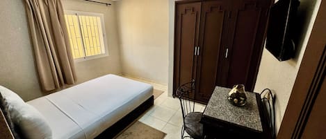 A compact bedroom with a bed, a desk, and a chair. There is a window with curtains, a wardrobe, and a television mounted on the wall.