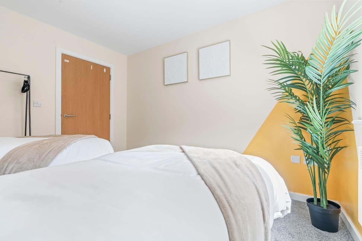 A hotel room with two beds, a door, a wall-mounted hairdryer, and a potted plant.