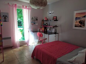 Ein Schlafzimmer mit einem Bett, einem Schreibtisch, einem Stuhl, einem Fenster mit Vorhängen sowie einer Wand mit Bildern und einem Kalender.