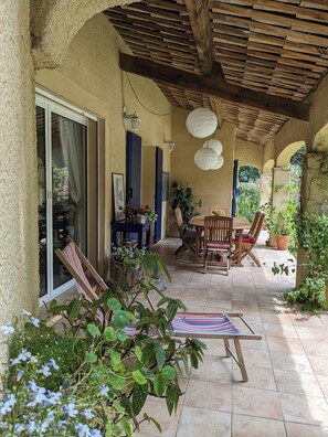 Eine überdachte Terrasse mit Tisch und Stühlen, ein gestreifter Stuhl und Kübelpflanzen.