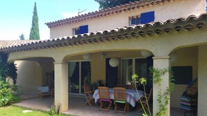 Une terrasse couverte avec une table et des chaises, un jardin avec de la pelouse et des plantes, et un bâtiment avec un toit en tuiles.