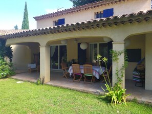 Eine überdachte Terrasse mit Tisch und Stühlen, ein Garten mit Rasen und Pflanzen und ein Gebäude mit Ziegeldach.