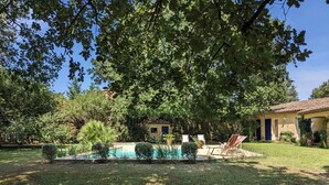 Une piscine entourée d’une pelouse bien entretenue, une maison avec un toit en tuiles et un grand arbre qui offre de l’ombre.