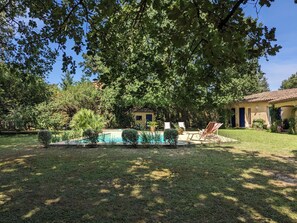 Ein Pool, umgeben von einer gepflegten Rasenfläche, ein Haus mit Ziegeldach und ein großer Baum, der Schatten spendet.