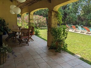 Una veranda coperta con tavolo e sedie, un'area piscina con lettini e un giardino con piante fiorite.