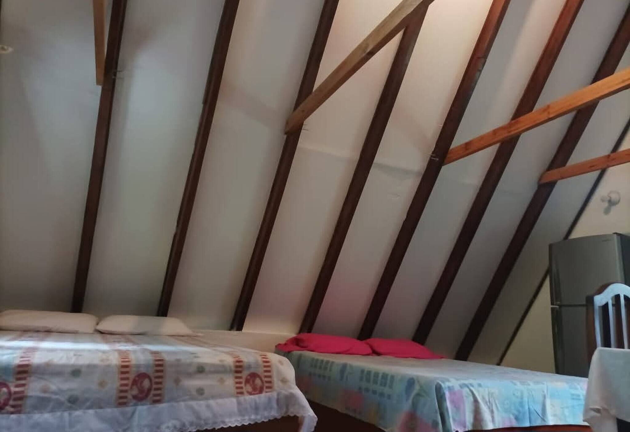 Una habitación con techo inclinado, vigas de madera, una cama con colcha estampada, una mesita con un objeto rojo encima y un altavoz.