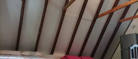A room with a sloped ceiling, wooden beams, a bed with a patterned bedspread, a small table with a red object on it, and a speaker.
