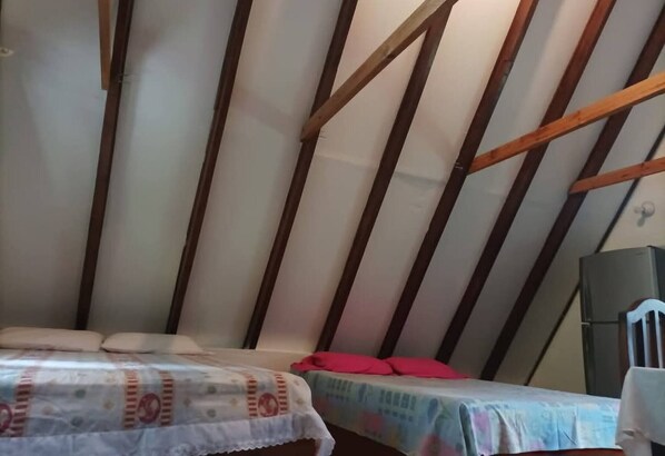 A room with a sloped ceiling, wooden beams, a bed with a patterned bedspread, a small table with a red object on it, and a speaker.