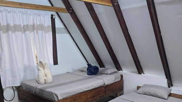 Un dormitorio pequeño tipo buhardilla con dos camas de madera, una ventana con cortinas blancas y suelo de baldosas.