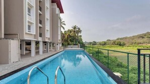 A swimming pool adjacent to a building with balconies, surrounded by greenery and a hilly landscape. - 2bhk With Pool nr Candolim Beach Palms By Umiya (Candolim)