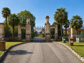 A gated entrance with a paved road leading through it, surrounded by palm trees and stone walls. - Elegant Lakeview Apartment for 6 (Germignaga)