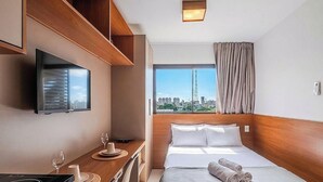Quarto de hotel com uma cama, televisão na parede, janela com vista para a cidade e mesa com um copo e uma xícara.