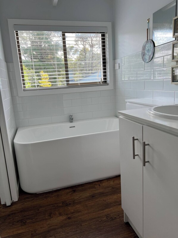 Un baño moderno con una bañera independiente, muebles blancos y un ventanal con persianas.