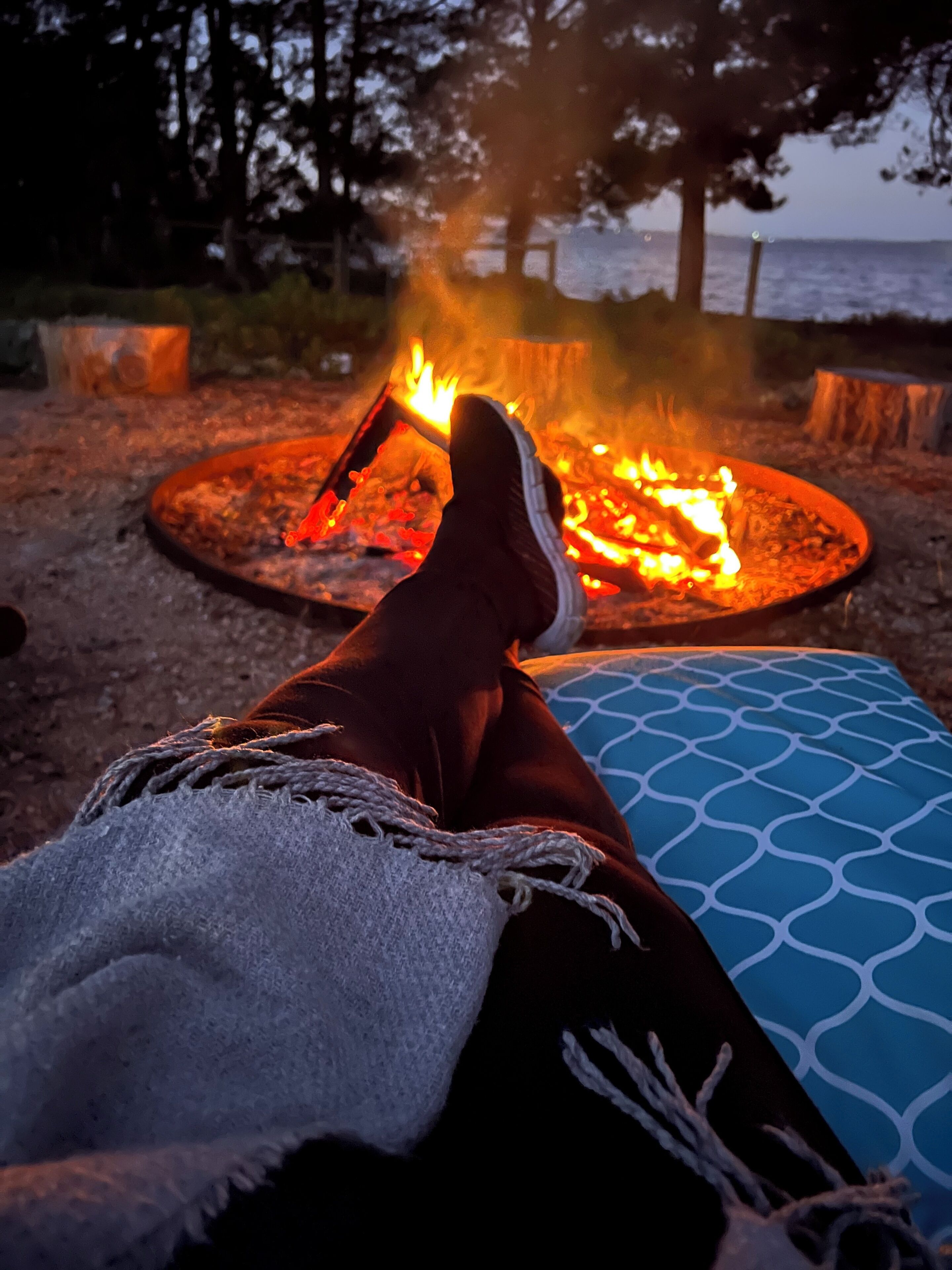A person relaxing by a campfire with a blanket and a blue patterned cushion.