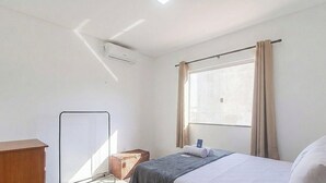 A hotel room with a bed, a bedside table, a small table, a window with curtains, and a ceiling light. - Apt. in Condo w/ Pool in Porto Seguro Maa0004 (Porto Seguro)
