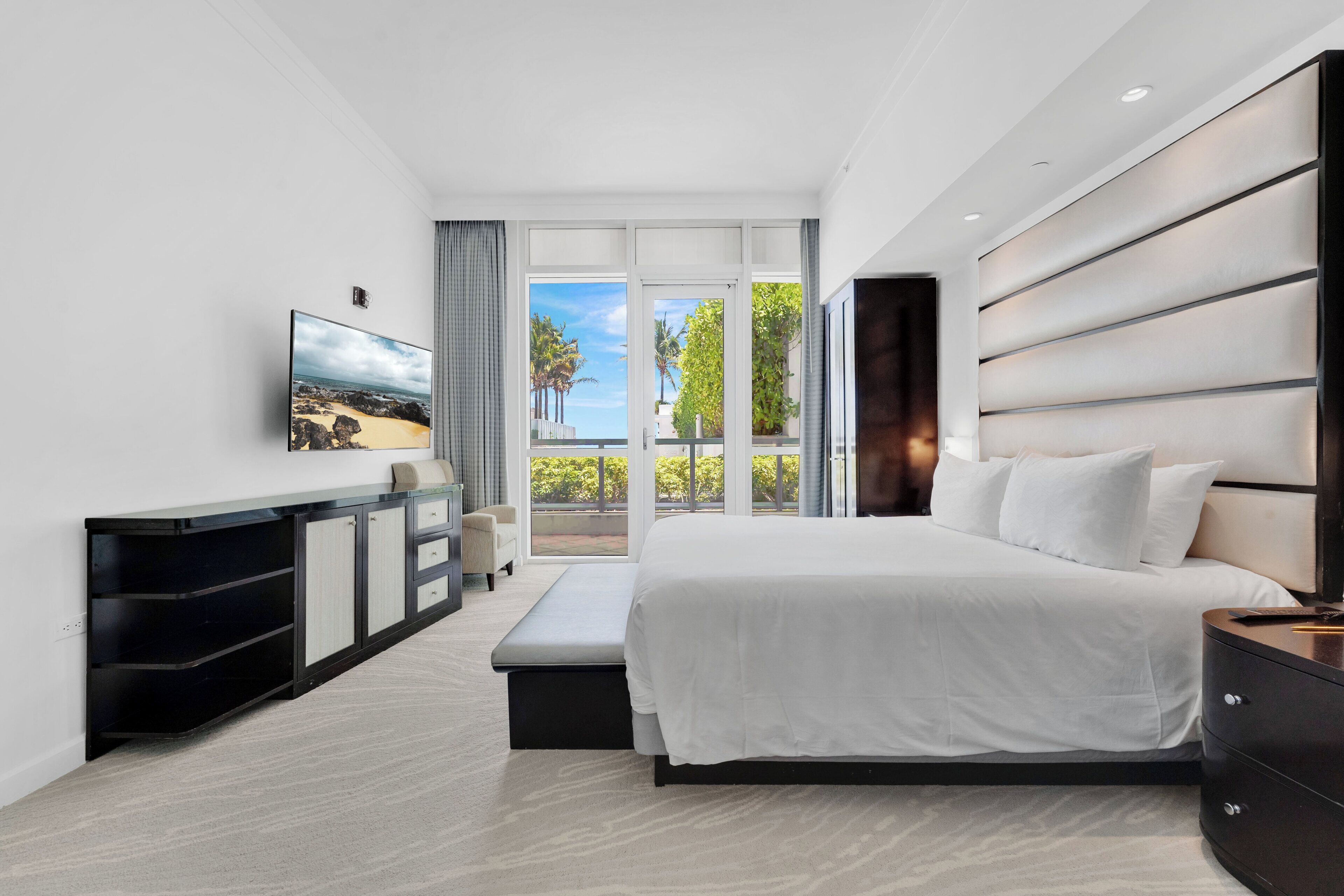 A modern hotel room with a large bed, a TV, a seating area, and a balcony with a view of the beach.